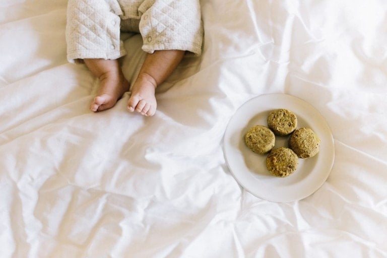 A baby in light-colored quilted clothing sits on a white bed. Next to the baby, a white plate holds four round bread rolls and hints at what could be a delicious lactation cookie recipe. The baby's legs and feet are visible, but their upper body is not in the frame.