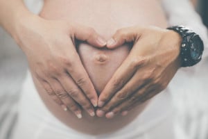 Two hands gently form a heart shape over the belly of a pregnant woman. The hands belong to a person wearing a black watch, and the woman's belly button is visible. The photograph focuses on the connection and tenderness between the individuals, symbolizing positive affirmations for pregnancy.