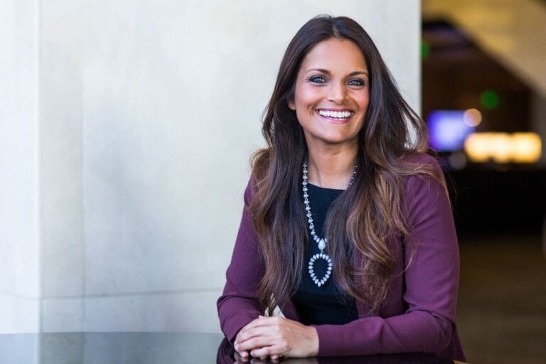 Dr. Shefali, with long brown hair, is sitting at a table, smiling. She is wearing a maroon blazer, a black top, and a necklace with a heart-shaped pendant. The background appears to be a well-lit indoor space with a light-colored wall and blurred elements.
