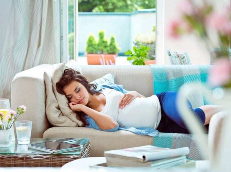 A pregnant woman experiencing pregnancy fatigue rests on a beige couch, lying on her side with one hand supporting her head and the other on her belly. She is dressed in a white shirt and blue pants. The room is bright with natural light and has a vase of flowers and a glass of water on a nearby table.