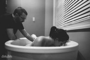 A black-and-white photo showing a man supporting a woman who is leaning forward in a bathtub. The woman, appearing to be in the transition labor phase, has her head down and seems to be in labor. The scene takes place in a room with a window that has blinds.