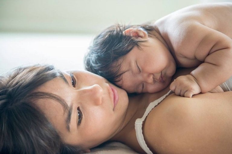 A woman lies on her back, looking off into the distance with a neutral expression. An infant rests on her chest, asleep with its head snuggled into her shoulder. The background is softly blurred, capturing the tender postpartum moment between mother and child.