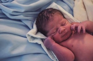 A newborn baby wrapped in a white towel lies on a bed with light blue sheets. The baby's eyes are closed, its tiny hands near its face. With dark hair and a calm expression, this serene moment reminds us of the things not to do during labor—such as forgetting to prepare for post-birth tranquility.