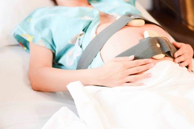 A pregnant person lies on a hospital bed with a fetal monitoring device strapped to their exposed belly, receiving essential birth tips. The person wears a hospital gown and rests one hand on the blanket covering their lower body.