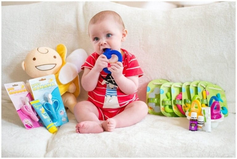 A baby wearing a red striped outfit sits on a white blanket, holding a blue teething ring. Surrounding the baby are various teething products and other baby care items, including a plush toy, teething toothbrushes, pacifiers, and baby oil.