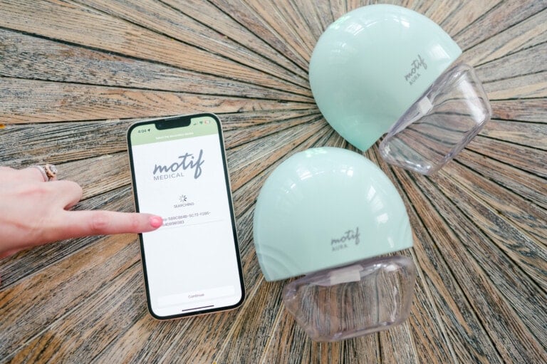 A person uses a finger to point at a smartphone screen displaying the text "motif medical" and "SEARCHING." Next to the phone, on a wooden surface, are two green motif Aura breast pump devices.