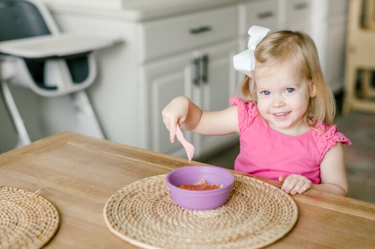 A young child with blonde hair and a white bow is sitting at a wooden table. She is wearing a pink sleeveless top and holding a pink spoon, eating yogurt from a purple bowl. In the background, there is a high chair and white cabinets.