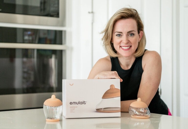 A person with shoulder-length hair is smiling while seated at a kitchen counter. In front of them is a box labeled "emulait" with two baby bottles on either side, perfect for parents who combine breastfeeding and bottle-feeding. The background features a microwave and other kitchen appliances.