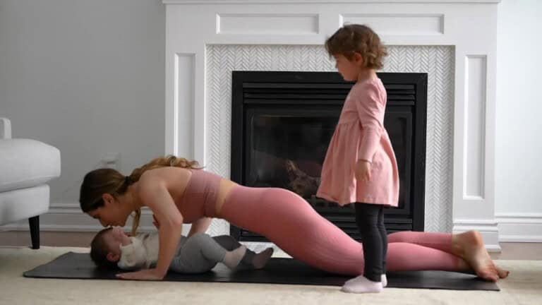 A woman is doing a push-up while looking at a baby lying beneath her. A young girl in a pink dress stands nearby. They are in a living room with a fireplace in the background.