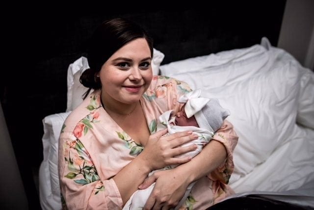 A woman wearing a floral robe is sitting on a bed, holding a newborn baby wrapped in a blanket at a birth center. The woman is smiling and looking at the camera. The baby has a bow on its head, and white pillows are visible in the background.