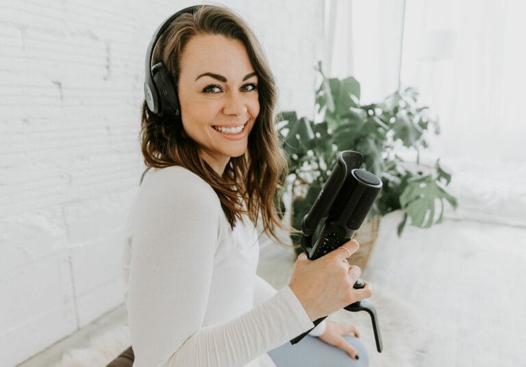 A person wearing headphones is smiling at the camera while holding a microphone. The person has long brown hair and is dressed in a white long-sleeved shirt. In the bright, airy room behind them, where mindfulness activities for kids are often practiced, there is a plant enhancing the serene ambiance.