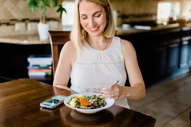 A woman with blonde hair, dressed in a white sleeveless top, sits at a wooden table holding a fork and smiling at a bowl of salad. Her smartphone is placed on the table next to the bowl. The background includes a kitchen area with a plant and stacked books, reflecting her focus on nutrition in pregnancy.