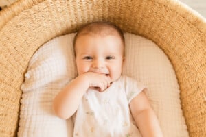 A baby is lying in a woven bassinet. The baby is wearing a light-colored onesie with small designs on it and is smiling with one hand near their mouth. As you consider cute boy names, this sweet moment could inspire you—perhaps something as comforting as the bassinet’s soft, cushioned lining.