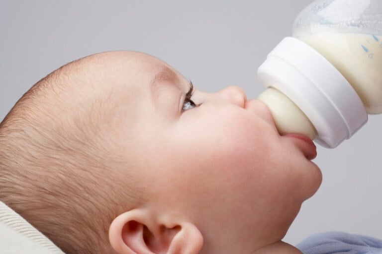 A close-up of an infant lying down while feeding from a baby bottle. The baby's head is turned to the side, and their mouth is latched onto the bottle's nipple, drinking milk, ensuring they are not overfeeding. The background is a plain light gray.
