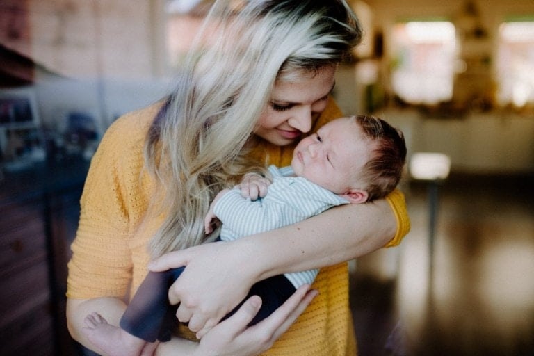 A woman with long blonde hair wearing a yellow sweater holds a baby dressed in a light blue and white striped outfit. They are indoors with a blurry background, and the woman is gently nuzzling the baby's nose, illustrating pure joy—perfect for showcasing gifts for new moms.