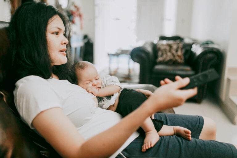 A woman sits on a couch, holding a remote control in her right hand and a sleeping baby on her lap. She wears a white t-shirt and dark shorts. The living room in the background has another couch and various household items, as she considers shows to binge-watch on maternity leave.