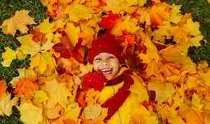 A child is lying on the ground covered in colorful autumn leaves, smiling widely. Engaging in a favorite fall activity, the child is wearing a red beanie, red scarf, and a yellow jacket, enjoying everything fall has to offer.