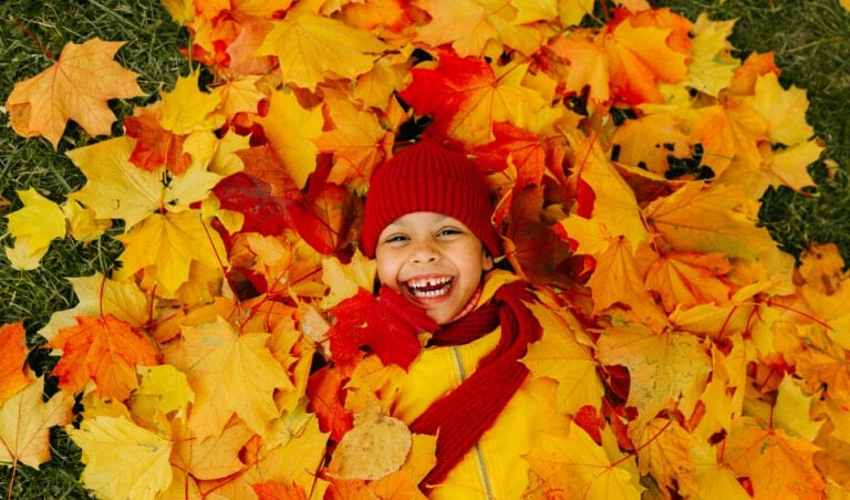 A child is lying on the ground covered in colorful autumn leaves, smiling widely. Engaging in a favorite fall activity, the child is wearing a red beanie, red scarf, and a yellow jacket, enjoying everything fall has to offer.