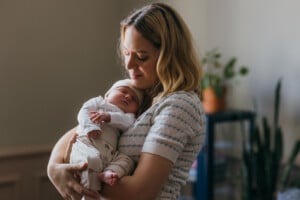 A woman is holding a sleeping baby in her arms. The woman has blonde hair and is wearing a light-colored striped top. The baby, possibly named after popular Welsh boy names, is dressed in light-colored clothing and is wearing a hat. There are plants and a piece of furniture in the blurred background.