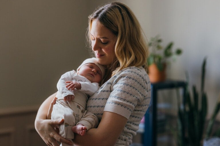 A woman is holding a sleeping baby in her arms. The woman has blonde hair and is wearing a light-colored striped top. The baby, possibly named after popular Welsh boy names, is dressed in light-colored clothing and is wearing a hat. There are plants and a piece of furniture in the blurred background.