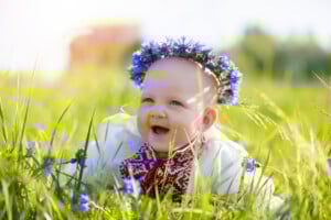 A baby wearing an embroidered white shirt and a purple flower crown is lying on the grass in a field. The baby, one of many who might bear beautiful Slavic names, is smiling, and a few purple flowers are visible around them. The background includes blurred greenery and sunlight.