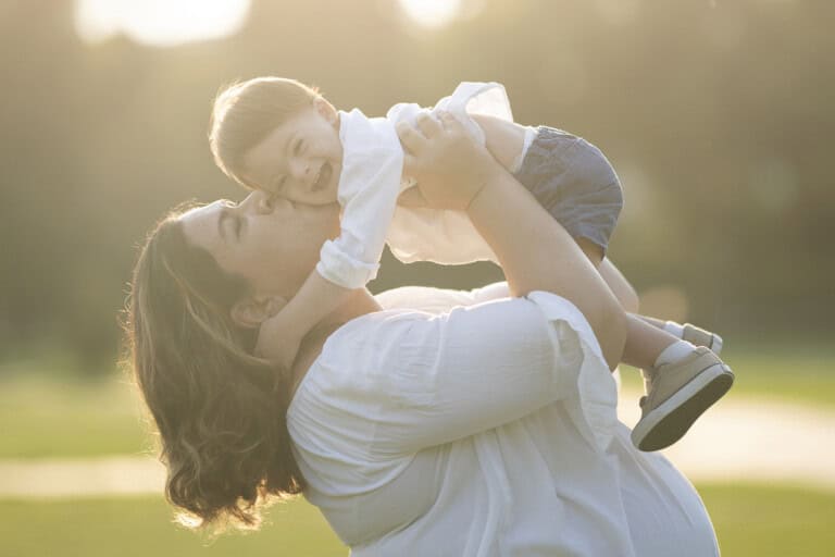 Young mother is kissing her little son with down syndrome. They are standing on the grass. She holds up her smiling 19-month-old in an outdoor setting, where the bright sunlight creates a soft, warm glow around them. The woman wears a white shirt, and the baby is in a white top and blue shorts with small shoes.