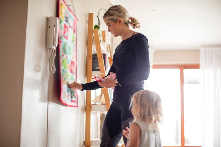 A woman dressed in black holds a toy next to a young child with blonde hair. They are looking at a colorful board with visual supports on the wall in a bright room, featuring a wooden ladder shelf and large windows in the background.