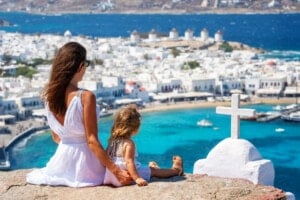 A woman and child sit on a rock overlooking a coastal town with white buildings, windmills, and turquoise water. The woman wears a white dress and sunglasses, while the child has on a dress and sandals. A white cross is visible in the foreground. The scene emanates the charm of Greek names like Alexios or Eleni.