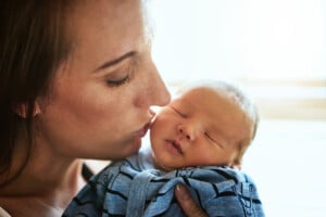 A person, appearing to be a mother, gently holds a newborn baby close to her face. The baby is sleeping, swaddled in a blue blanket, while the person gazes affectionately at the Irish boy. Natural light illuminates the scene from the background.