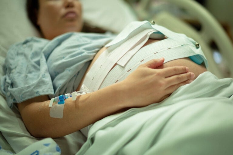 A pregnant woman lies in a hospital bed, wearing a hospital gown. A fetal monitoring belt is wrapped around her abdomen, and an IV is inserted into her arm. Her left hand rests on her belly as she awaits treatment for chorioamnionitis. The background shows part of the hospital bed and bedding.