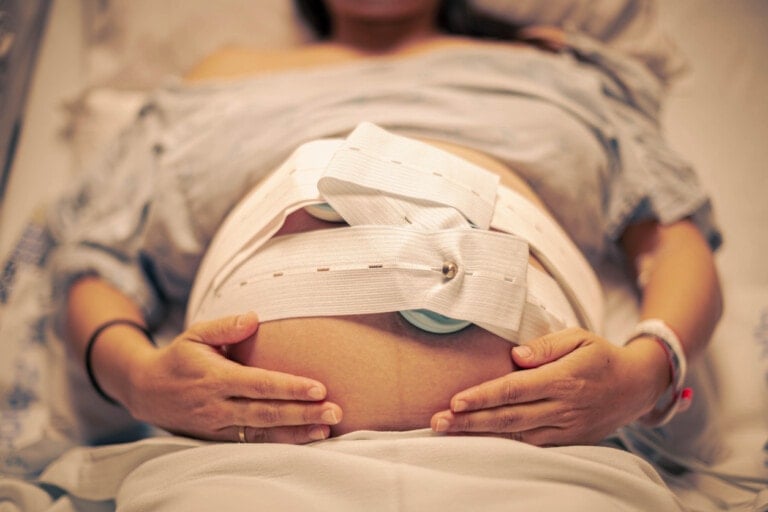 A pregnant woman lies in a hospital bed, her hands gently resting on her abdomen. Fetal monitoring belts track the baby's heartbeat and contractions, vigilant for any pregnancy complications. A medical wristband encircles her wrist, and she is covered with a light-colored blanket.
