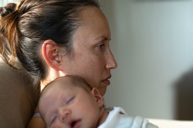 A woman with her hair tied back is sitting and looking away, appearing deep in thought. She is holding a sleeping baby close to her chest, possibly reflecting on postpartum mood disorders. Both are indoors with soft lighting from a window casting gentle shadows on the woman's face.