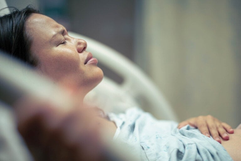 A woman lying in a hospital bed appears to be in pain, with her eyes closed and her face contorted. She is wearing a hospital gown, and her hand is resting on her abdomen, indicating the aftermath of a 3rd degree perineal tear. The background is out of focus, highlighting the woman as the main subject.