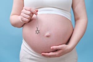 A pregnant person in a white outfit holds a Star of David necklace with one hand while the other hand rests on their bare belly. The background is light blue, evoking thoughts of possible Hebrew names for their child.