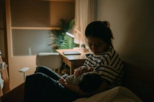 A woman seated in a dimly lit room is breastfeeding a baby, perhaps engaging in dream feeding. She is wearing a striped shirt and her hair is tied back. In the background, there is a desk with a lamp, potted plant, and various items on top. The room appears calm and serene.