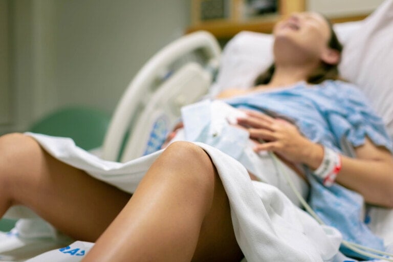 A woman is lying in a hospital bed wearing a hospital gown. She appears to be in labor, her legs bent and partially covered by a white sheet. Medical equipment and monitors are visible beside the bed, dispelling any myths about labor and birth through the vigilance of modern healthcare.