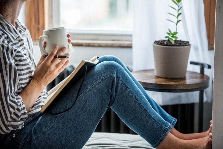 A person sits by a window holding a mug and a pen, with a notebook resting on their lap, resembling planners for moms. They are wearing a striped shirt and blue jeans. A potted plant is placed on a wooden side table next to them.