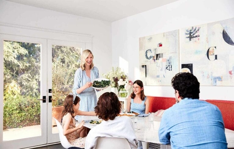 A group of five people are gathered around a dining table in a bright room. Catherine McCord, standing next to the table, is serving food. The room has large windows with a view of trees and abstract art on the walls. The table is adorned with plates of delicious food and drinks.