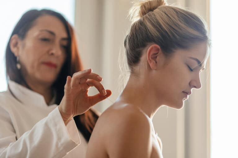 A young woman is seated while another woman, in a white coat, administers acupuncture by inserting a thin needle into her back near the shoulder. The seated woman has her eyes closed and hair tied up. The background is softly lit, suggesting a calm environment.