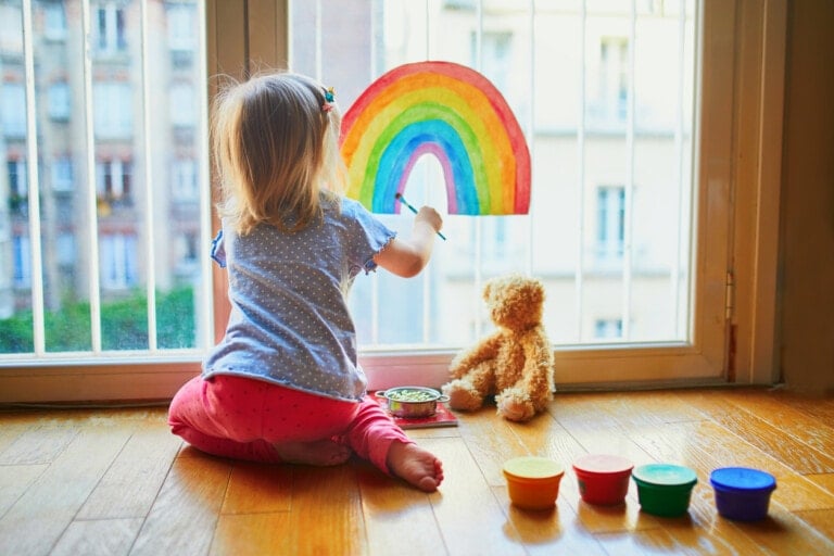 A young child with blonde hair sits on the floor, facing a large window. The child is painting a colorful rainbow on a piece of paper taped to the window—a perfect example of fun indoor activities for kids. A teddy bear and several containers of paint are nearby on the wooden floor.