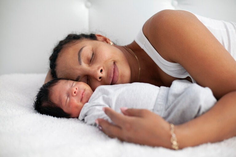 A woman and a baby are sleeping closely together on a white bed. The woman, facing the camera with a content expression despite the pressure on moms, lies next to her baby, who is peacefully resting on its back. Both are dressed in light-colored clothing.