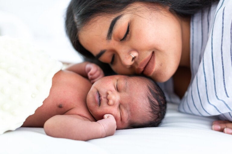 A woman with long dark hair, wearing a striped shirt, rests her head next to a sleeping newborn baby. The mama's serene expression complements the peaceful look on her baby's face as they lie on a white blanket with a textured yellow blanket partially covering them.