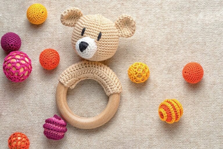 A delightful knitted teddy bear rattle with a wooden ring is surrounded by colorful crocheted balls on a textured beige fabric surface, perfect for those looking for homemade baby gifts.