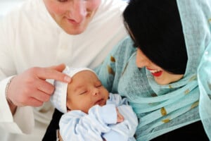 A newborn baby is held by a smiling woman wearing a light blue headscarf. A man dressed in a white shirt touches the baby's head gently while looking down at the sleeping infant with a smile. The baby, dressed in a blue striped outfit, may be destined for one of many beautiful Arabic names for boys.