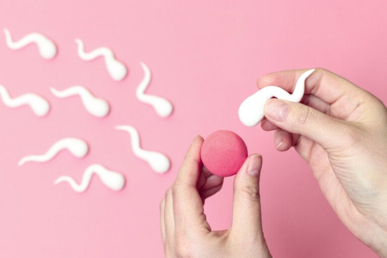 A person holds a white sperm-shaped object in their right hand and a small red ball in their left. Several more sperm-shaped objects are arranged diagonally on a pink background, evoking themes of understanding infertility.