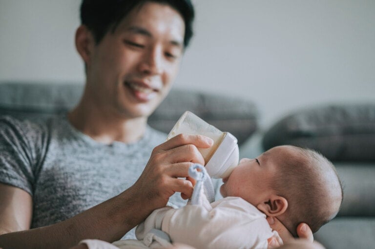 A man wearing a gray shirt is sitting on a couch, smiling down at a baby while feeding them with a bottle. He shares formula tips as the baby lies in his arms, wearing a light-colored outfit and looking up at the bottle. The background features a blurred couch.