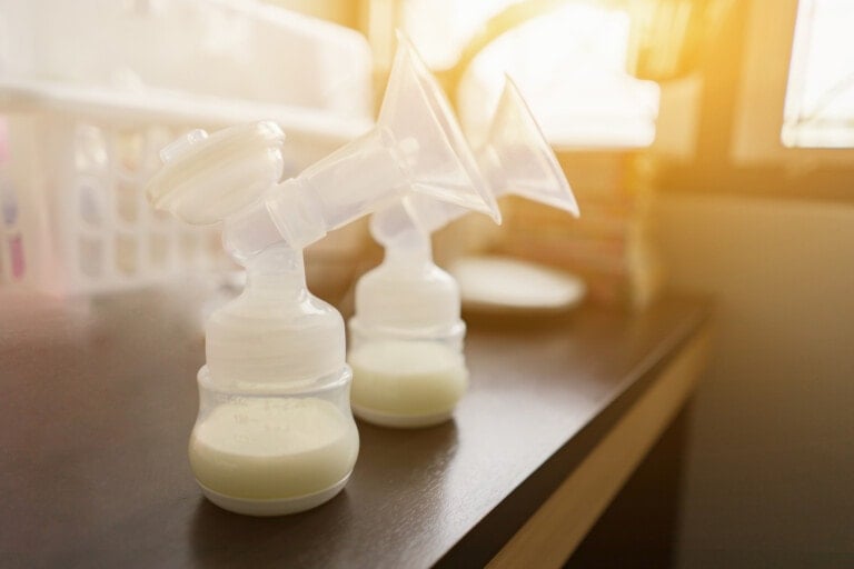 A double electric breast pump with filled milk bottles is placed on a dark wooden surface. Sunlight streams in from a nearby window, illuminating the plastic parts of the pump, capturing the essence of every time you pump. In the background, soft focus containers and items rest gently on a shelf.