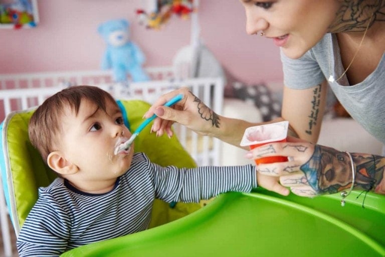 A tattooed adult is feeding a baby in a high chair with a spoon and a container of probiotic yogurt. The baby, wearing a striped shirt, has food on their face. A white crib and blue teddy bear are seen in the background.