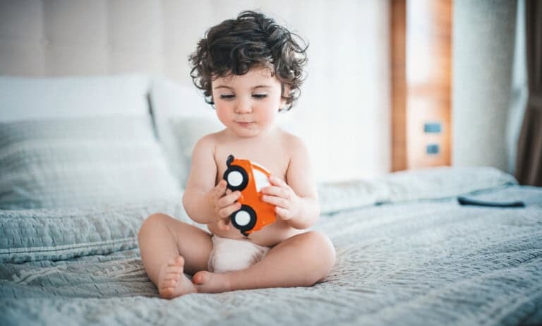 A 21-month-old boy with curly hair sits on a bed, wearing only a diaper. The toddler is holding and examining a small red toy car with black wheels. The background reveals a softly lit room, hinting at the comforting environment key to maintaining a consistent sleep schedule for toddlers this age.