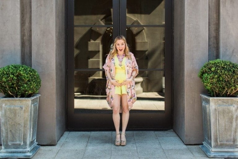 A woman in a yellow dress and floral kimono, who appears to be in the second trimester of pregnancy, stands between two potted plants in front of a large glass door. She has a surprised expression on her face with her hands placed on her stomach. The background includes a stone building facade.